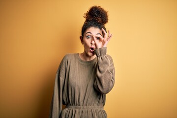 Fototapeta na wymiar Young beautiful brunette woman with curly hair and piercing wearing casual dress doing ok gesture shocked with surprised face, eye looking through fingers. Unbelieving expression.