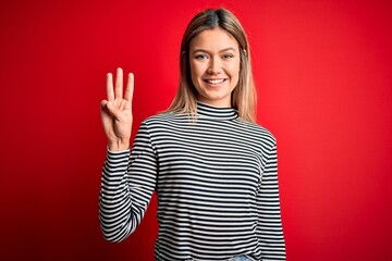 Young beautiful blonde woman wearing casual striped sweater over red isolated background showing and pointing up with fingers number three while smiling confident and happy.