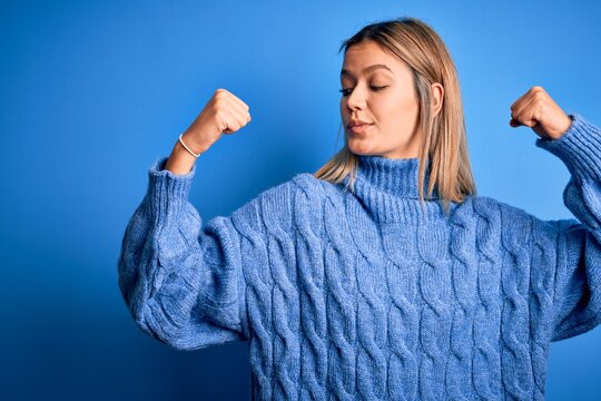 Young beautiful blonde woman wearing winter wool sweater over blue isolated background showing arms muscles smiling proud. Fitness concept.