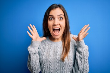 Beautiful young woman wearing casual wool sweater standing over blue isolated background celebrating crazy and amazed for success with arms raised and open eyes screaming excited. Winner concept