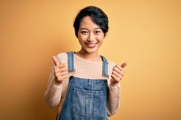 Young beautiful asian girl wearing casual denim overalls over isolated yellow background success sign doing positive gesture with hand, thumbs up smiling and happy. Cheerful expression and winner