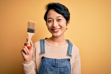 Young beautiful chinese woman painting holding paint brush over isolated yellow background with a happy face standing and smiling with a confident smile showing teeth