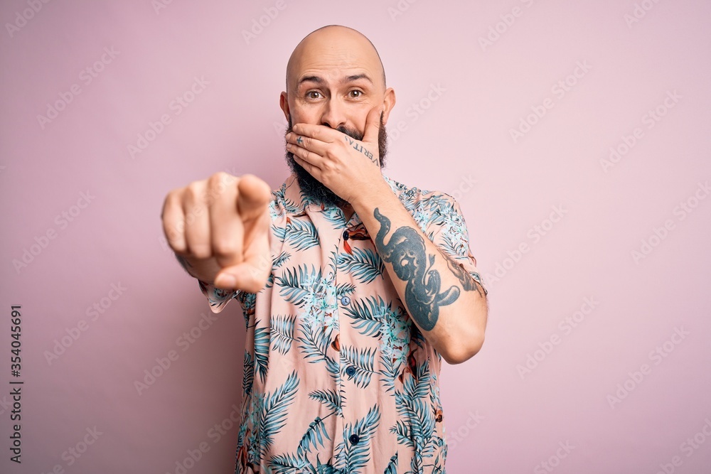 Sticker handsome bald man with beard and tattoo wearing casual floral shirt over pink background laughing at