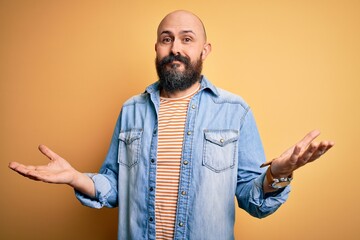 Handsome bald man with beard wearing casual denim jacket and striped t-shirt clueless and confused expression with arms and hands raised. Doubt concept.