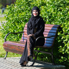 A beautiful young muslim woman is sitting on a bench. Islamic girl smiles broadly, resting on a...