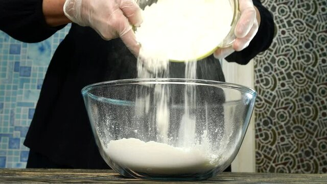 Female cook prepares dough for baking. Pour wheat flour into transparent glass bowl, add kefir, salt, olive machelo and egg. Close-up.