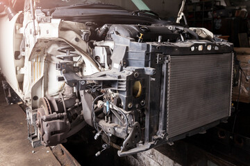 The front part of the car after an accident in a car repair service with a disassembled hood and metal grille and various parts in the garage box.
