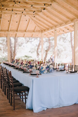 Outdoor banquet table under the wooden roof of the gazebo. Rack and cutlery, purple glasses. Floral arrangement of violet, blue, black, silver flowers. Black banquet chairs. On the table is a blue tab