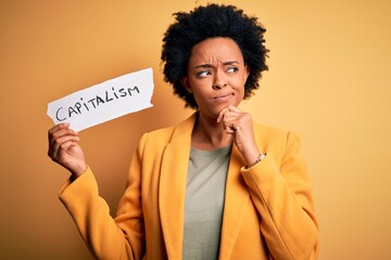 African American afro businesswoman with curly hair holding paper with capitalism message serious...
