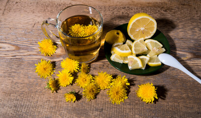 Mug of tea with yellow dandelions, a green saucer, lemon, sugar on a wooden table