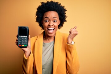 Young African American afro woman with curly hair holding dataphone payment device screaming proud...