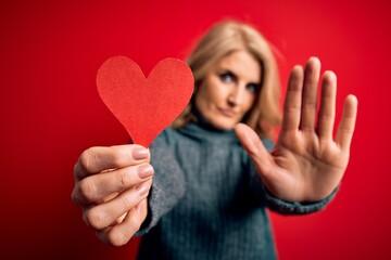 Middle age beautiful blonde romantic woman holding red paper heart celebrating valentine day with open hand doing stop sign with serious and confident expression, defense gesture