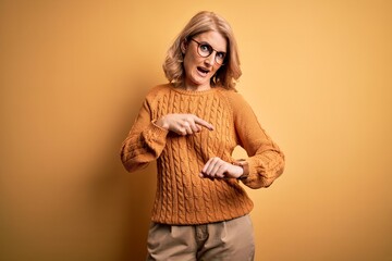 Middle age beautiful blonde woman wearing casual sweater and glasses over yellow background In hurry pointing to watch time, impatience, upset and angry for deadline delay