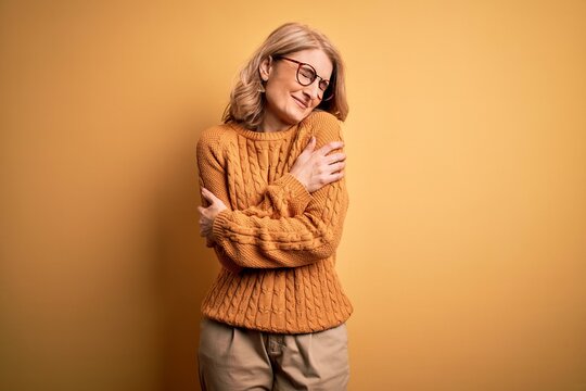 Middle age beautiful blonde woman wearing casual sweater and glasses over yellow background Hugging oneself happy and positive, smiling confident. Self love and self care