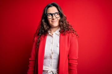 Young beautiful woman with curly hair wearing jacket and glasses over red background smiling looking to the side and staring away thinking.