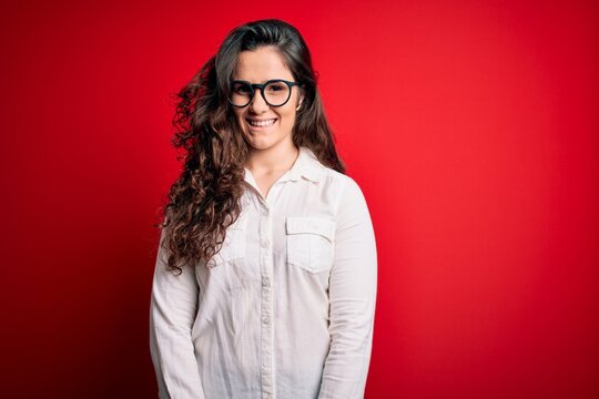 Young beautiful woman with curly hair wearing shirt and glasses over red background with a happy and cool smile on face. Lucky person.