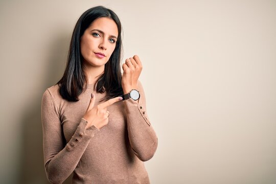 Young brunette woman with blue eyes wearing casual sweater over isolated white background In hurry pointing to watch time, impatience, looking at the camera with relaxed expression