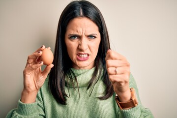 Young brunette woman with blue eyes holding fresh raw egg over isolated background annoyed and...