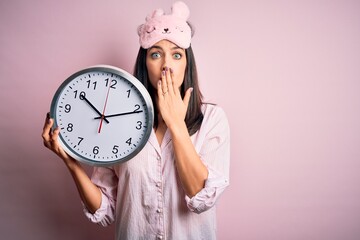 Young brunette woman with blue eyes wearing pajama holding big clock at night time cover mouth with...