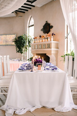 Presidium, banquet table with a white tablecloth. Plates and cutlery, purple glasses, napkins, candles in glass candlesticks. Bouquet of flowers, asters, roses, carnations, eustomas.
