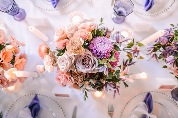 Presidium, banquet table with a white tablecloth. Plates and cutlery, purple glasses, napkins, candles in glass candlesticks. Bouquet of flowers, asters, roses, carnations, eustomas.