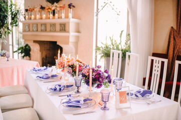 Presidium, banquet table with a white tablecloth. Plates and cutlery, purple glasses, napkins, candles in glass candlesticks. Bouquet of flowers, asters, roses, carnations, eustomas.