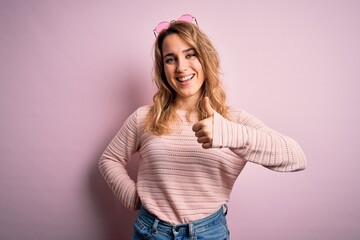 Young beautiful blonde woman wearing casual sweater and sunglasses over pink background doing happy thumbs up gesture with hand. Approving expression looking at the camera showing success.
