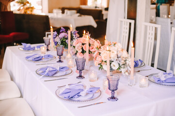 Presidium, banquet table with a white tablecloth. Plates and cutlery, purple glasses, napkins, candles in glass candlesticks. Bouquet of flowers, asters, roses, carnations, eustomas.