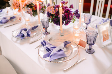 Presidium, banquet table with a white tablecloth. Plates and cutlery, purple glasses, napkins, candles in glass candlesticks. Bouquet of flowers, asters, roses, carnations, eustomas.
