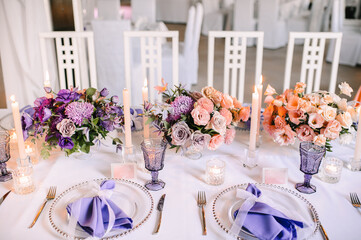 Presidium, banquet table with a white tablecloth. Plates and cutlery, purple glasses, napkins, candles in glass candlesticks. Bouquet of flowers, asters, roses, carnations, eustomas.