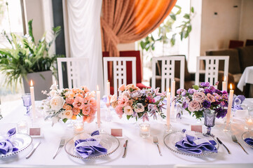 Presidium, banquet table with a white tablecloth. Plates and cutlery, purple glasses, napkins, candles in glass candlesticks. Bouquet of flowers, asters, roses, carnations, eustomas.