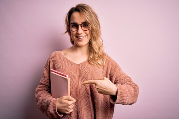 Young beautiful blonde woman wearing glasses holding notebook over pink background very happy pointing with hand and finger