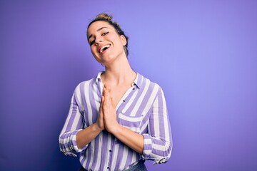 Young beautiful blonde woman wearing casual striped shirt standing over purple background praying with hands together asking for forgiveness smiling confident.