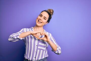 Young beautiful blonde woman wearing casual striped shirt standing over purple background smiling in love doing heart symbol shape with hands. Romantic concept.