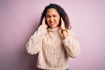 Young beautiful woman with curly hair wearing casual sweater standing over pink background covering ears with fingers with annoyed expression for the noise of loud music. Deaf concept.