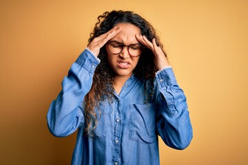 Beautiful woman with curly hair wearing casual denim shirt and glasses over yellow background with hand on head for pain in head because stress. Suffering migraine.