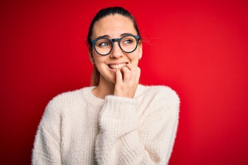 Beautiful blonde woman with blue eyes wearing sweater and glasses over red background looking stressed and nervous with hands on mouth biting nails. Anxiety problem.