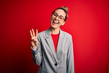 Young beautiful blonde businesswoman with blue eyes wearing glasses and jacket showing and pointing up with fingers number three while smiling confident and happy.
