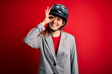 Young beautiful blonde motorcyclist woman wearing motorcycle helmet over red background doing ok gesture with hand smiling, eye looking through fingers with happy face.