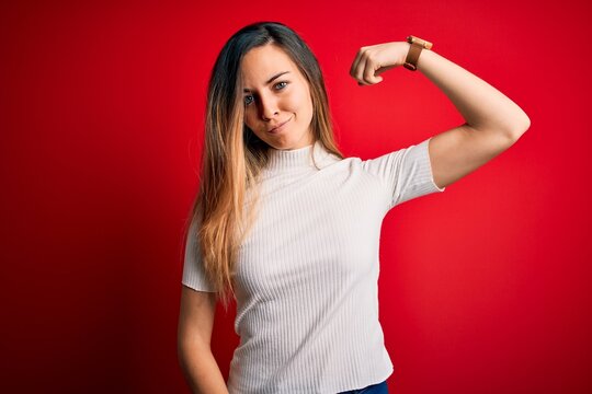 Beautiful blonde woman with blue eyes wearing casual white t-shirt over red background Strong person showing arm muscle, confident and proud of power