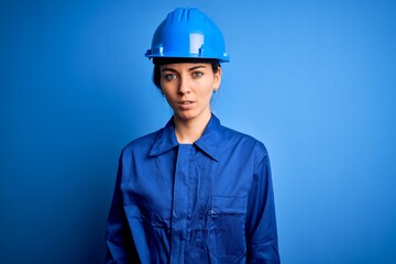 Young beautiful worker woman with blue eyes wearing security helmet and uniform with serious expression on face. Simple and natural looking at the camera.