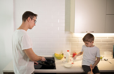 Boy son helps a young happy father prepare food in a bright kitchen