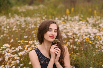 Young beautiful girl with closed eyes holds flowers in hands in chamomile field