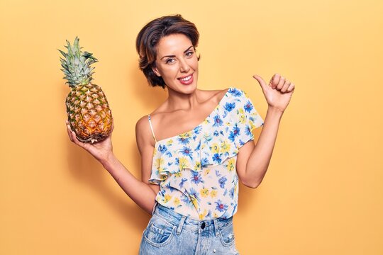 Young beautiful woman holding pineapple smiling happy and positive, thumb up doing excellent and approval sign