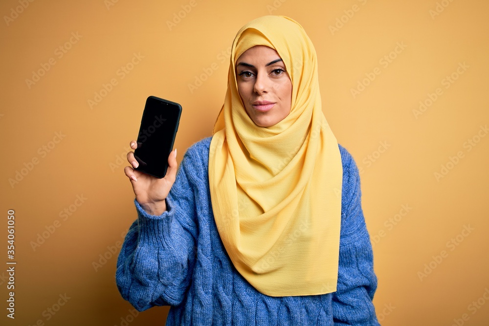 Poster young beautiful brunette arab woman wearing islamic hijab holding smartphone showing screen with a c