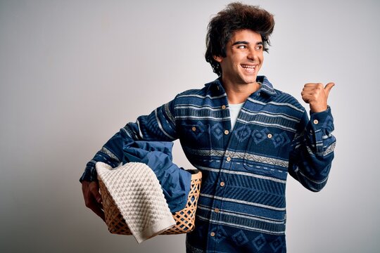 Young handsome man holding wicker basket with clothes over isolated white background pointing and showing with thumb up to the side with happy face smiling