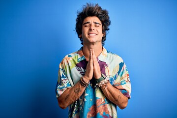 Young handsome man on vacation wearing summer shirt over isolated blue background begging and...