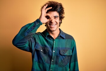 Young handsome man wearing casual shirt standing over isolated yellow background doing ok gesture with hand smiling, eye looking through fingers with happy face.