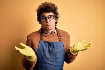 Young handsome cleaner man wearing apron and gloves over isolated yellow background clueless and...
