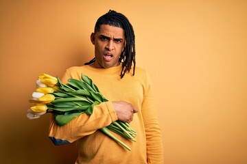 Young african american afro romantic man with dreadlocks holding bouquet of yellow tulips In shock face, looking skeptical and sarcastic, surprised with open mouth
